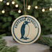 A round ornament with a penguin design, featuring the text "EMPOROR PENGUIN" on it, is displayed in front of a blurred background with Christmas lights.