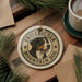A circular ornament featuring a mountain dog's head, with the text "ENTLEBUCKER MOUNTAIN DOG" printed on it, is displayed on a wooden surface alongside a cup of coffee and some pine branches.