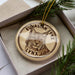 A circular ornament with a picture of a lynx on it, placed inside a box with pine branches.
