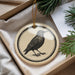 A decorative ornament featuring a black and white illustration of a bird is displayed in a box, surrounded by pine branches.