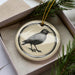 A ceramic ornament in a box, featuring a black and white illustration of a bird perched on a branch.