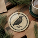 a round ceramic ornament with a black and white illustration of a bird, placed on a wooden surface next to a cup of coffee and some pine branches.
