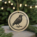 A round ornament featuring a black and white illustration of a bird is displayed in front of a blurred background of Christmas lights.