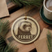 A round ornament featuring a ferret's face is displayed on a wooden surface, surrounded by pine branches and a cup of coffee.