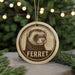 A round ornament featuring a ferret's face, with the word "FERRET" written below it, is displayed in front of a blurred Christmas tree.
