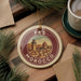 A round ornament with a cityscape design, featuring a tower and buildings, is displayed on a wooden surface alongside a cup of coffee and some pine branches.