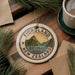 A circular ornament with the text "FIORDLAND NATIONAL PARK" and a mountain landscape design, placed on a wooden surface next to a cup of coffee and some pine branches.