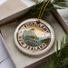 A decorative ornament in a box, featuring a circular design with text and a mountain landscape.