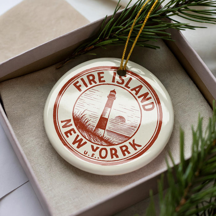 A white ornament with a red border and a lighthouse image, placed in a box with pine branches.