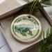 A round ornament with a green frog image and the text "FIRE-BELLED TOAD" printed on it, placed inside a box with pine branches.
