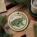 A round ornament featuring a green frog on a wooden surface, with the text "FIRE-BELLIED TOAD" printed on it.