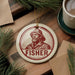 A round ornament featuring a man's portrait and the word "FISHER" in a red and white color scheme, placed on a wooden surface alongside pine branches and a cup of coffee.