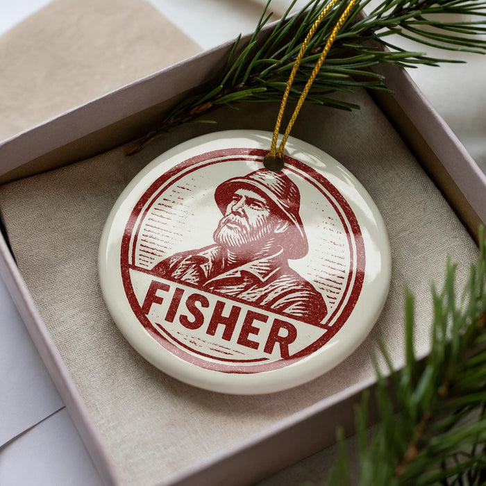 A round ornament with a portrait of a man wearing a hat and the word "FISHER" printed on it, placed in a box with pine branches.