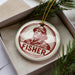 A round ornament with a portrait of a man wearing a hat and the word "FISHER" printed on it, placed in a box with pine branches.