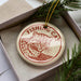 A round ornament with a cat image and the text "FISHING CAT" is displayed in a box, surrounded by pine branches.