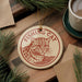 A round ornament with a cat image and the text "Fishing Cat" is displayed on a wooden surface, surrounded by pine branches and a cup of coffee.