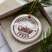 A white ceramic ornament with a maroon border and the text "FREDERICKSBURG TEXAS USA" printed on it, placed inside a box with pine branches.