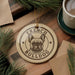 A circular ornament featuring a bulldog's face, with the text "FRENCH BULLDOG" printed on it, is displayed on a wooden surface alongside a cup of coffee and some pine branches.