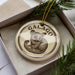 A circular ornament with a drawing of a small animal, likely a rodent or rodent-like creature, is displayed in a box with pine branches.