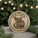 A circular ornament featuring a drawing of a small animal, likely a rodent or rodent-like creature, with the word "GALAGO" written around the edge. The ornament is hanging from a string, and there are pine branches visible in the background.