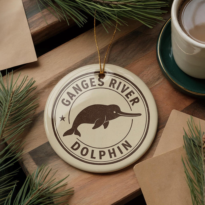 A circular ornament with the text "Ganges River Dolphin" and a graphic of a dolphin is displayed on a wooden surface, surrounded by pine branches and a cup of coffee.