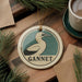 A round ornament featuring a bird, specifically a gannet, is displayed on a wooden surface alongside a cup of coffee and some papers.