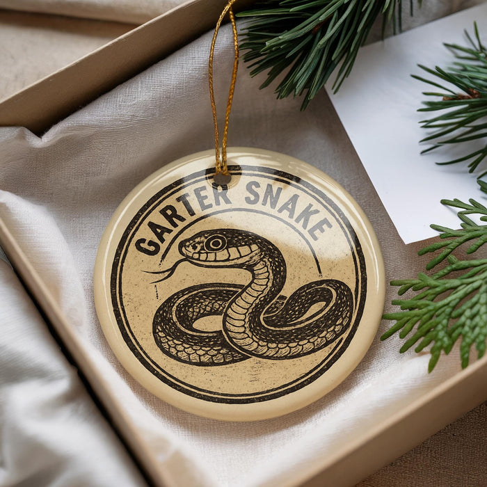 A round ornament with a snake design, hanging from a string, placed inside a box with pine branches.