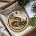 A round ornament with a snake design, hanging from a string, placed inside a box with pine branches.