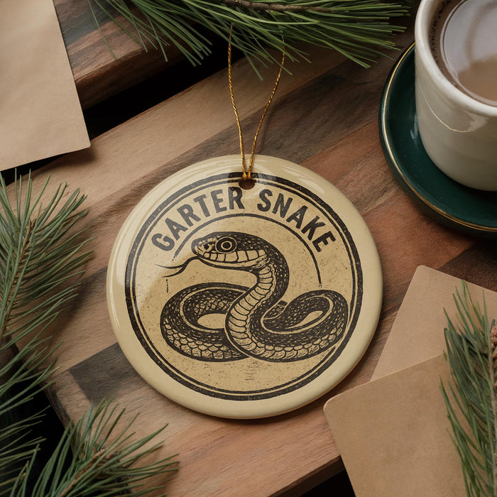 A round ornament with a snake design and the text "GARTER Snake" is displayed on a wooden surface, surrounded by pine branches and a cup of coffee.