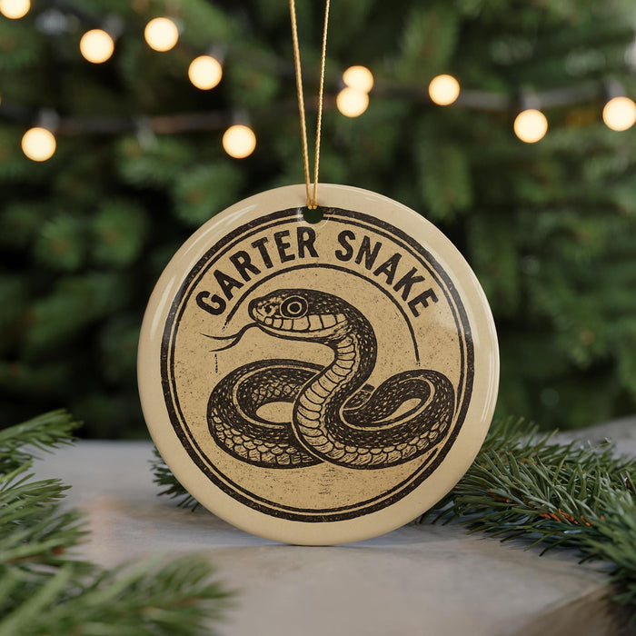 A round ornament with a snake design and the text "GARTER SNake" on it, hanging in front of a blurred Christmas tree with lights.