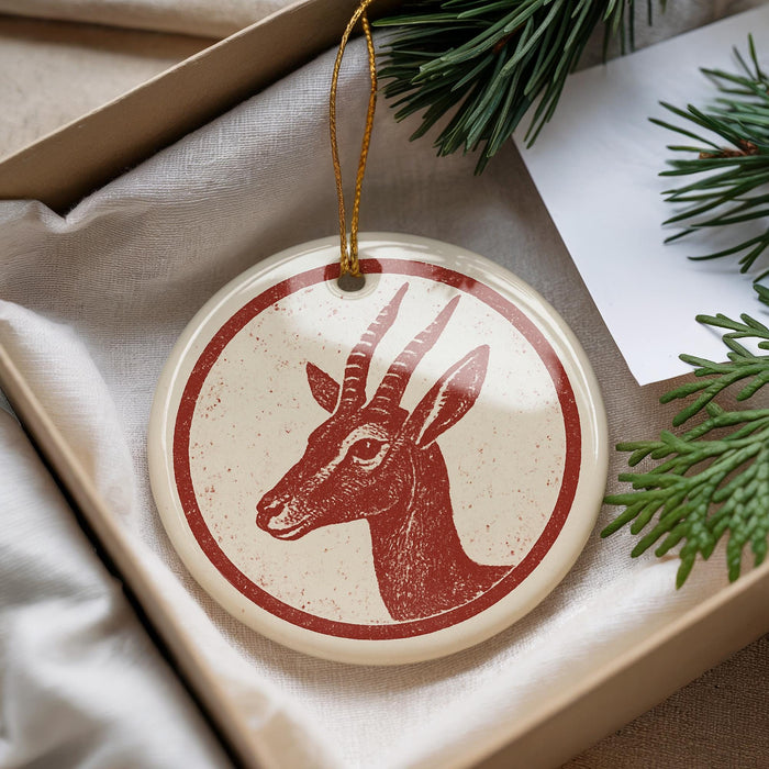 A round ornament with a red and white design featuring a deer head is displayed in a box, surrounded by pine branches.