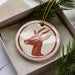 A ceramic ornament in the shape of a deer head is displayed in a box, surrounded by pine branches.