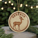 A circular ornament featuring a brown gazelle or deer with horns, with the word "Gereuk" written around the edge. The ornament is hanging from a string, and there are pine branches visible in the background.