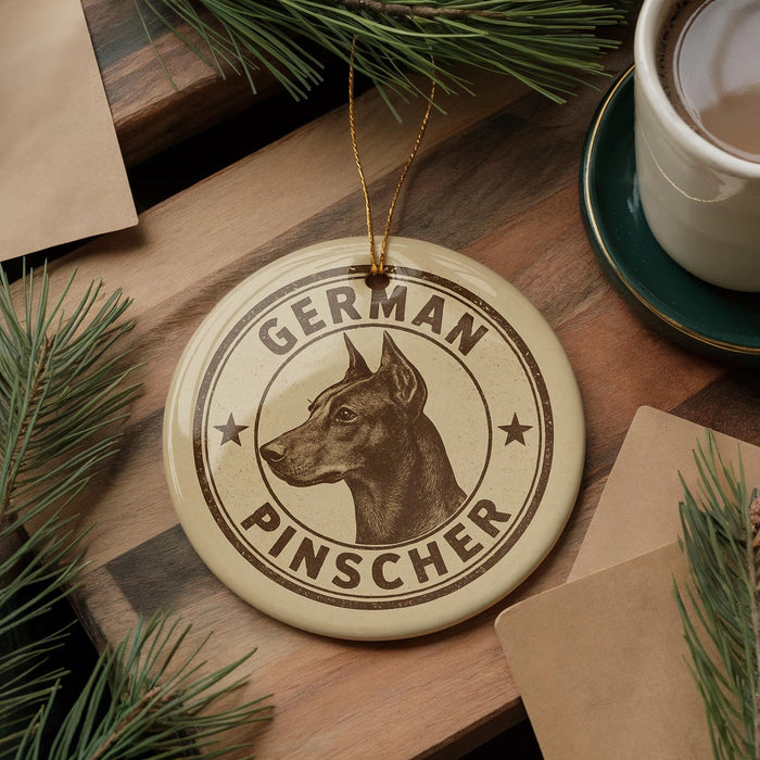 A round ornament with a dog's head and the text "GERMAN PINSCHER" is displayed on a wooden surface, surrounded by pine branches and a cup of coffee.
