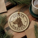 A round ornament with a dog's head and the text "GERMAN PINSCHER" is displayed on a wooden surface, surrounded by pine branches and a cup of coffee.