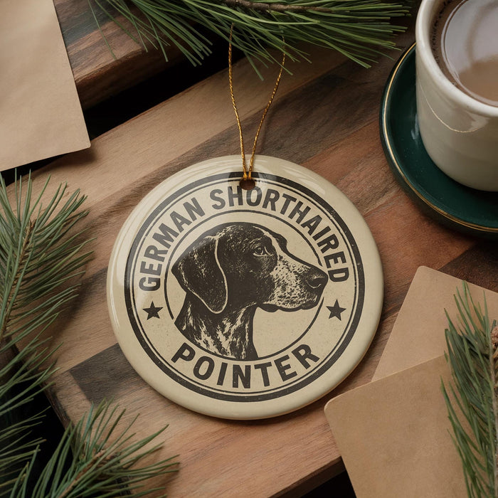 A circular ornament featuring a German Shorthaired Pointer dog, with the text "German Shorthaired Pointer" printed on it. The ornament is placed on a wooden surface, surrounded by pine branches and a cup of coffee.
