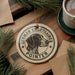 A circular ornament featuring a German Shorthaired Pointer dog, with the text "German Shorthaired Pointer" printed on it. The ornament is placed on a wooden surface, surrounded by pine branches and a cup of coffee.