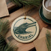 A ceramic ornament featuring a crocodile image and the word "GHARIAL" is displayed on a wooden surface, surrounded by pine branches and a cup of coffee.