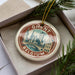A decorative ornament in a box, featuring a circular design with a cityscape and the text "Ghent, Belgium" prominently displayed.