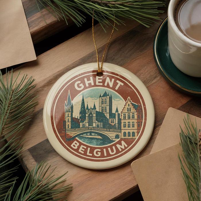 A circular ornament with a picture of a city skyline and the text "Ghent, Belgium" is displayed on a wooden surface, surrounded by pine branches and a cup of coffee.