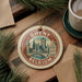 A circular ornament with a picture of a city skyline and the text "Ghent, Belgium" is displayed on a wooden surface, surrounded by pine branches and a cup of coffee.