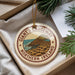A circular ornament with a design depicting a coastal landscape, featuring a rocky cliff and a body of water, is displayed in a box with pine branches.
