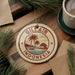 A round ornament with the text "GILI AIR INDONESIA" and a palm tree design, placed on a wooden surface next to a cup of coffee and some pine branches.