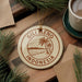 A circular ornament with the text "GILI MENO INDONESIA" printed on it, surrounded by pine branches and a cup of coffee on a wooden surface.