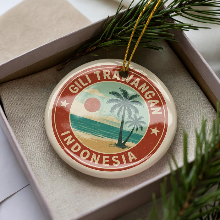 A circular ornament with a palm tree and beach scene, surrounded by a red border, is displayed in a box with pine branches.