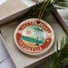 A circular ornament with a palm tree and beach scene, surrounded by a red border, is displayed in a box with pine branches.