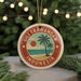 A circular ornament with a palm tree and beach scene, surrounded by a red border, hangs in front of a Christmas tree with twinkling lights.