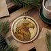 a round ornament with a detailed eagle head design, placed on a wooden surface alongside a cup of coffee and some pine branches.