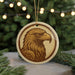 A circular ornament featuring a detailed illustration of an eagle's head is displayed against a blurred background of Christmas lights and pine branches.