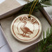A circular ornament with the word "GOOSE" printed on it, featuring a depiction of a goose. The ornament is placed inside a box, surrounded by pine branches.